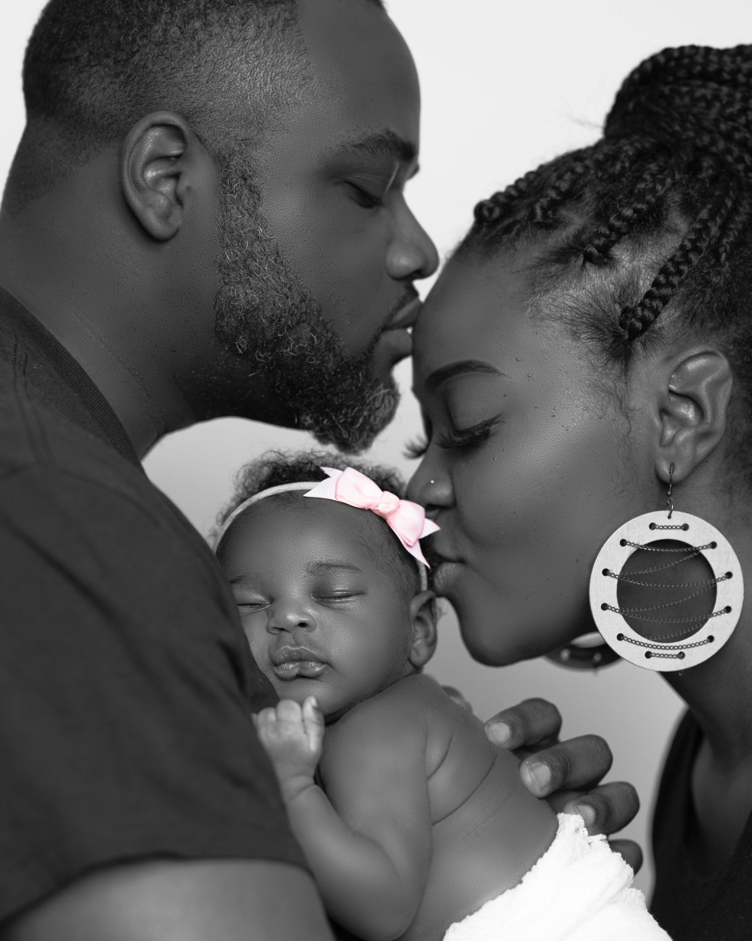A tender black and white image of parents embracing their sleeping newborn baby wearing a headband.