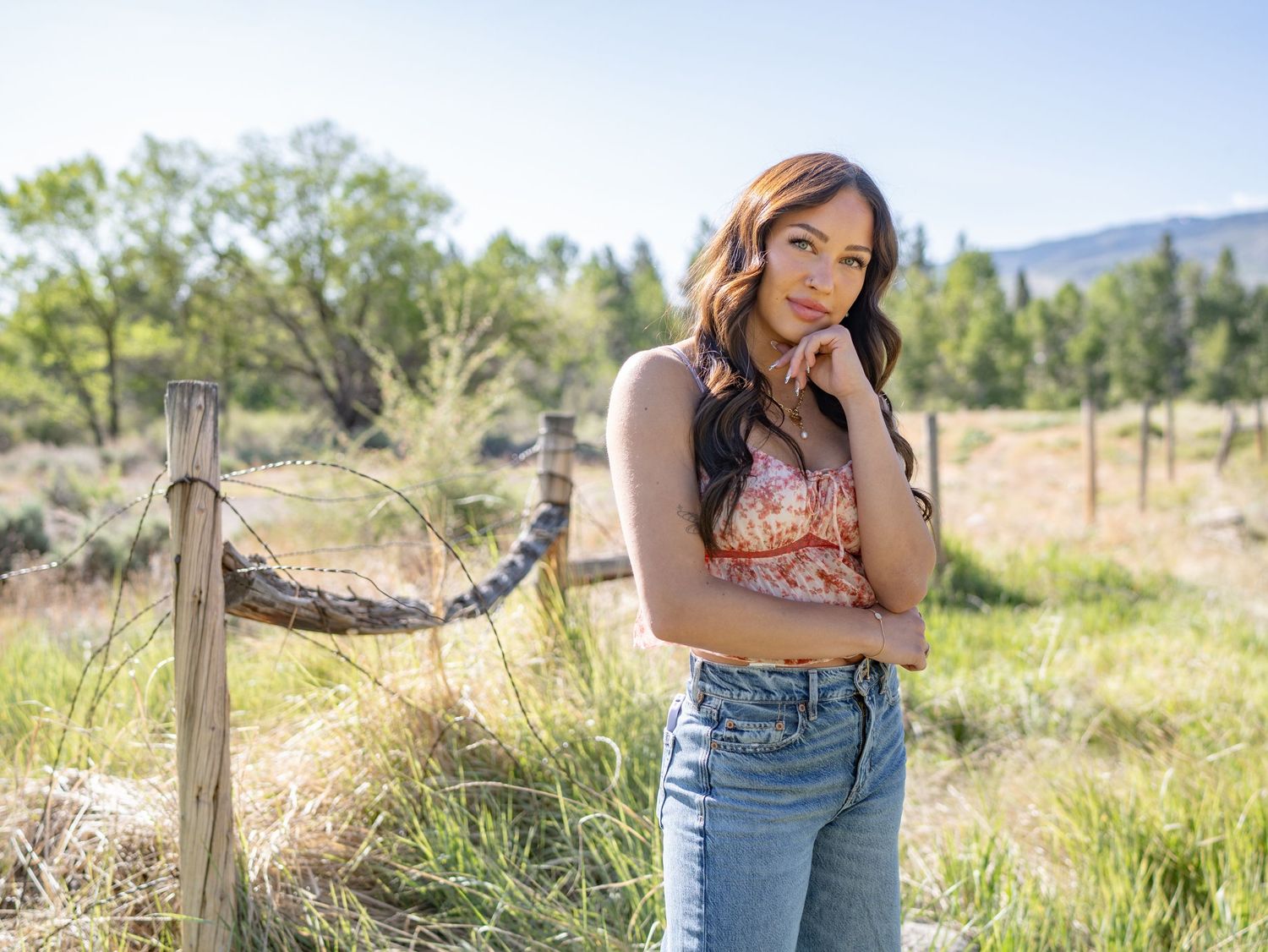 Senior in casual summer attire leaning against rustic wooden fence post in sunny rural field with mountains in background.