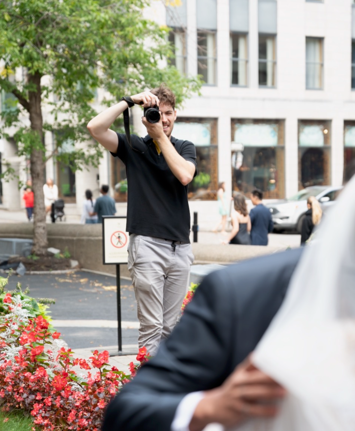 Urban photographer in black shirt taking pictures with camera near blooming red flowers and office buildings.