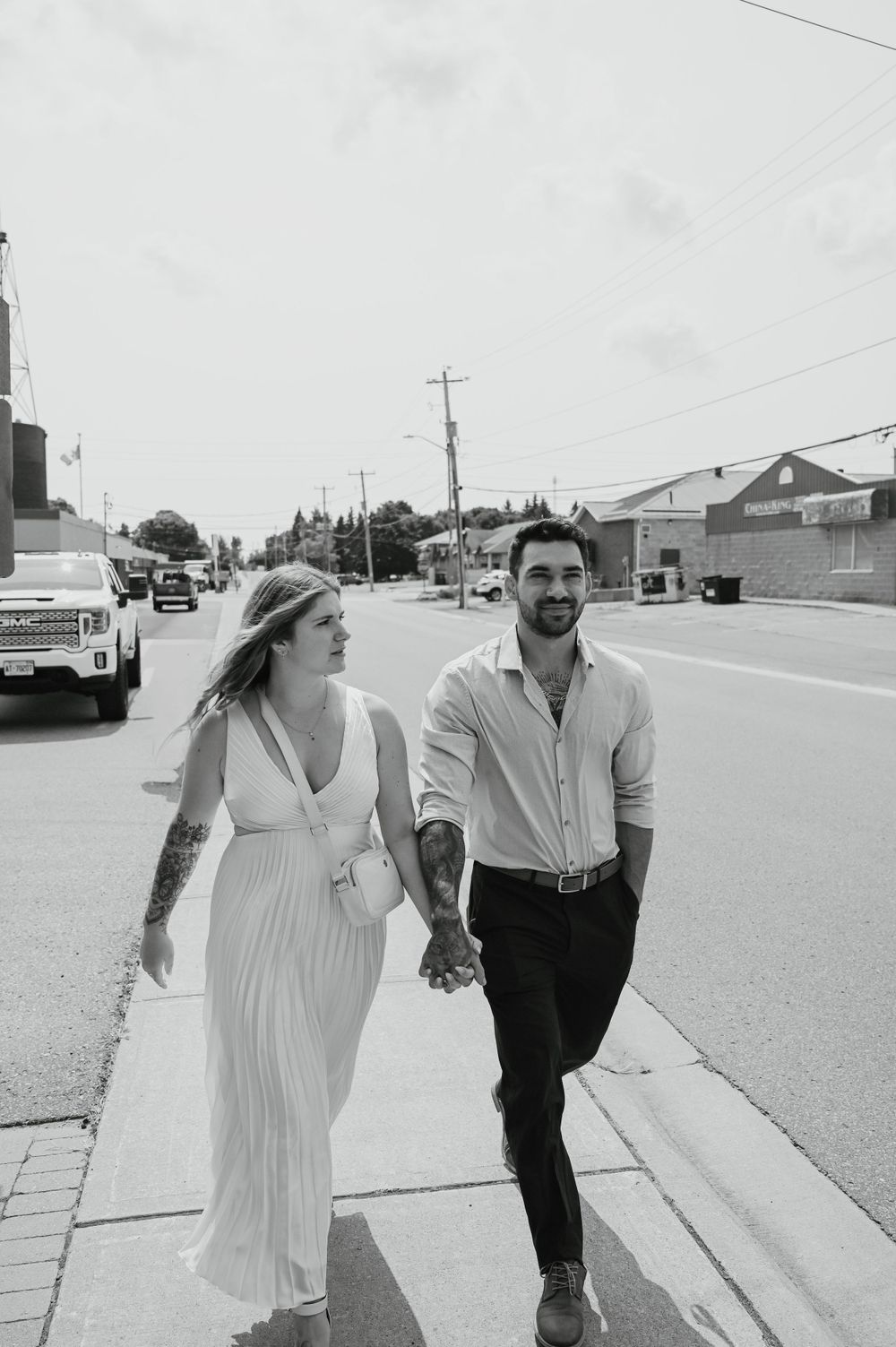 A couple wearing casual attire walks hand in hand down a suburban street on a sunny day captured in black and white.