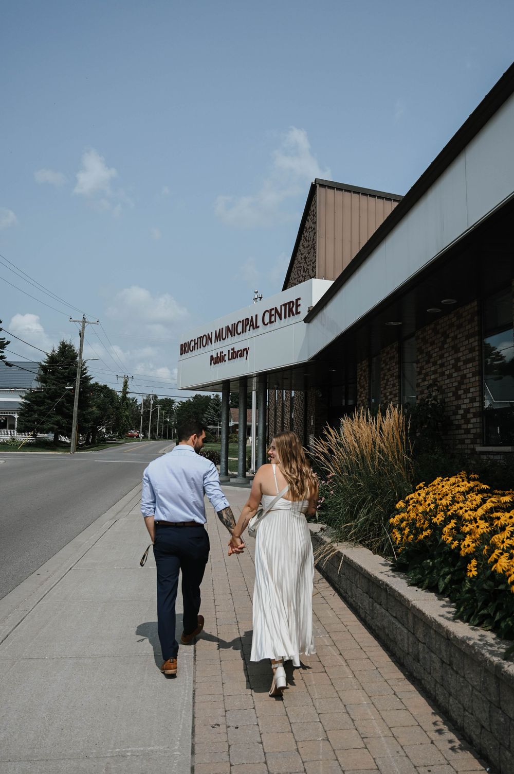 A couple walks hand in hand on a brick sidewalk past a medical centre with yellow flowers blooming in the garden.