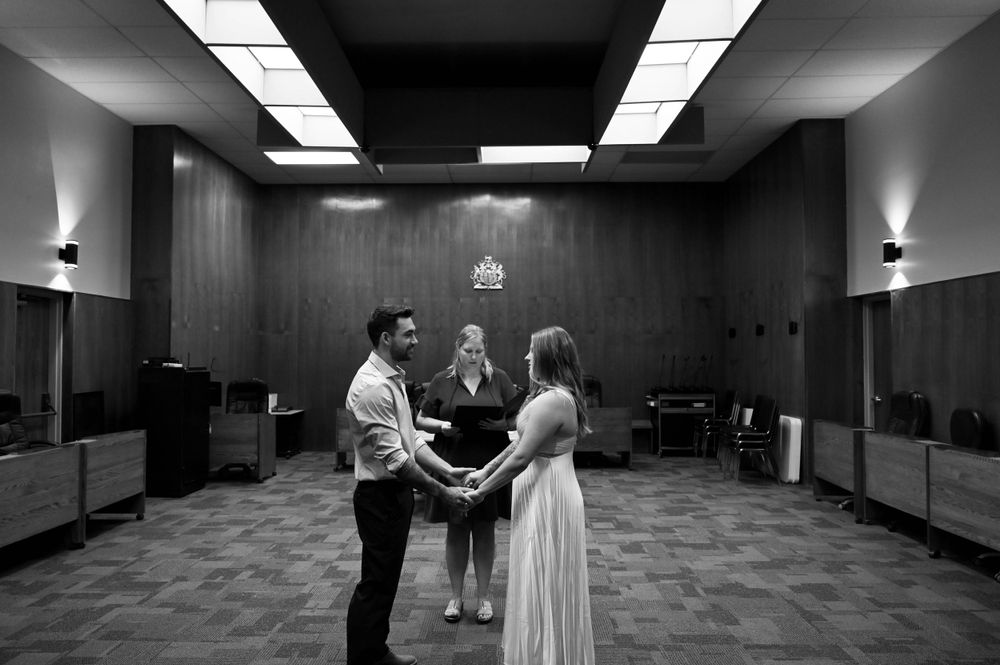 A couple stands together during what appears to be an intimate wedding ceremony in a black and white indoor setting.