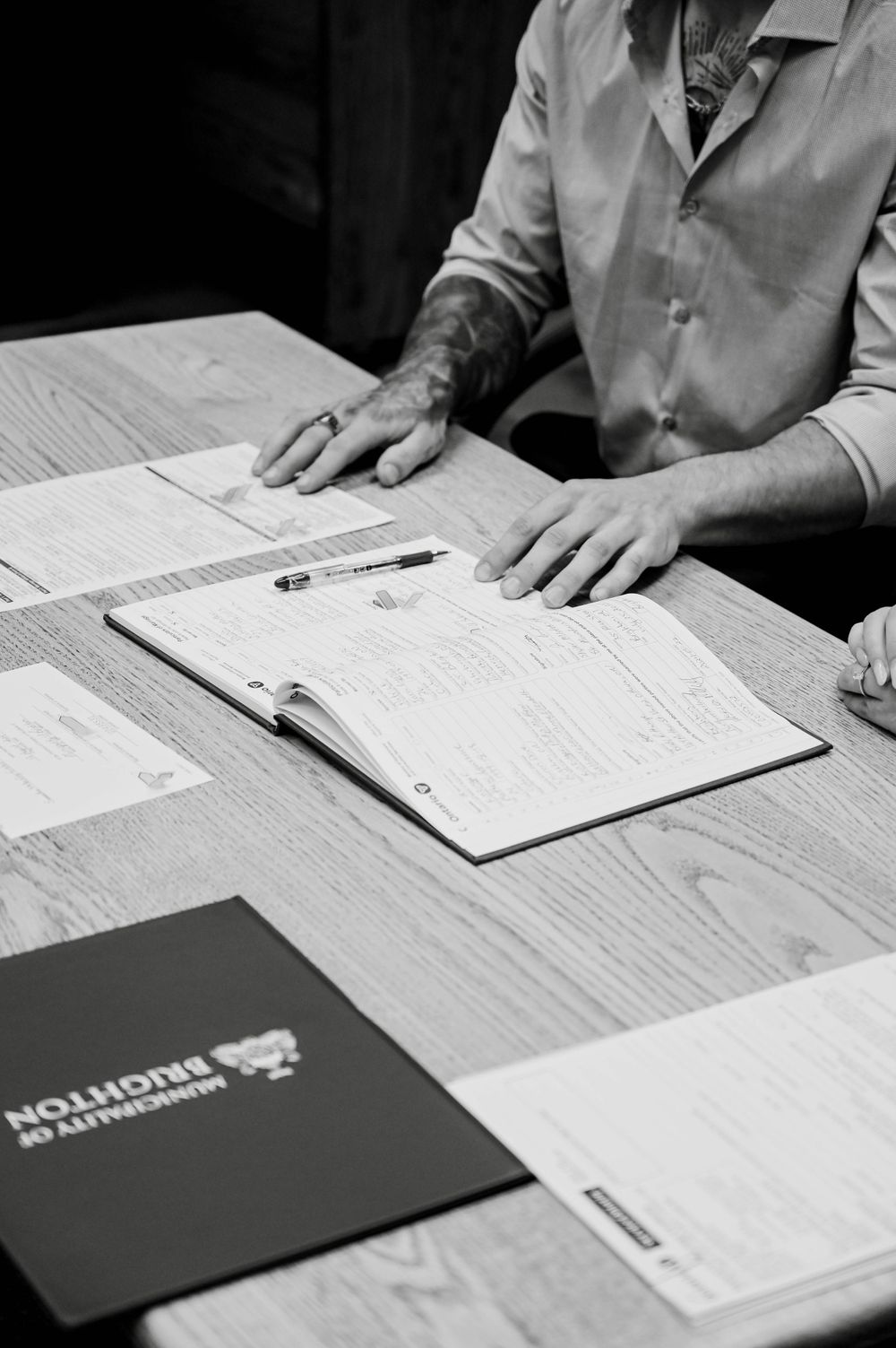 A person in a white coat reviews paperwork and documents on a wooden desk with a portfolio folder visible.