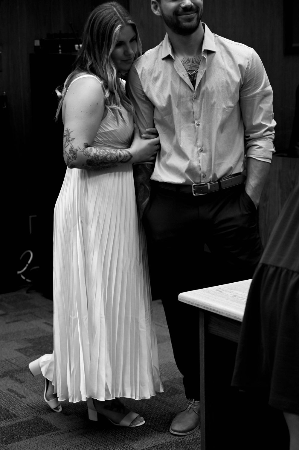 A black and white photograph shows a couple standing together, with the woman wearing a flowing white pleated dress.