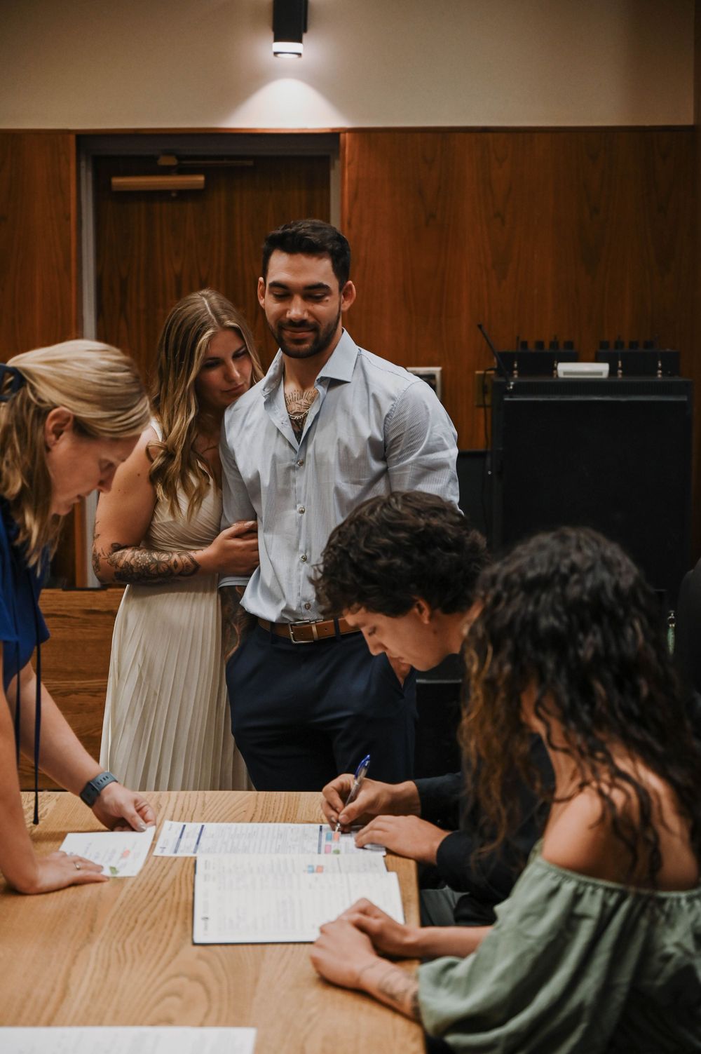 A couple and others gather around a table to sign documents in what appears to be an official setting.