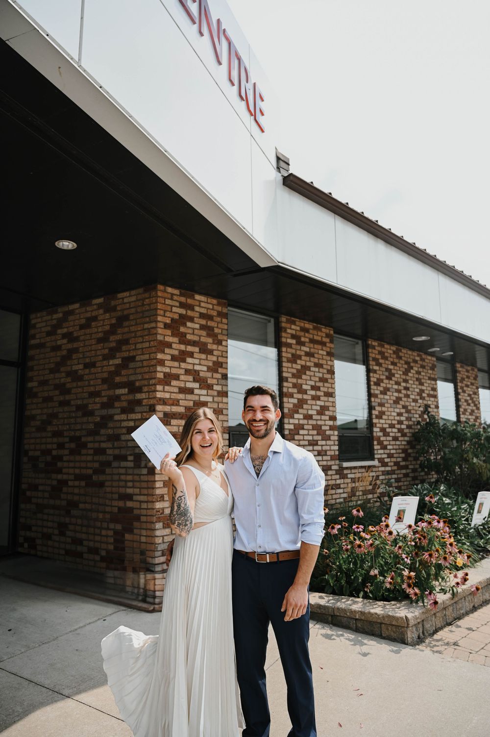 A couple stands outside a brick building in formal attire, with the person on the left holding up a document.