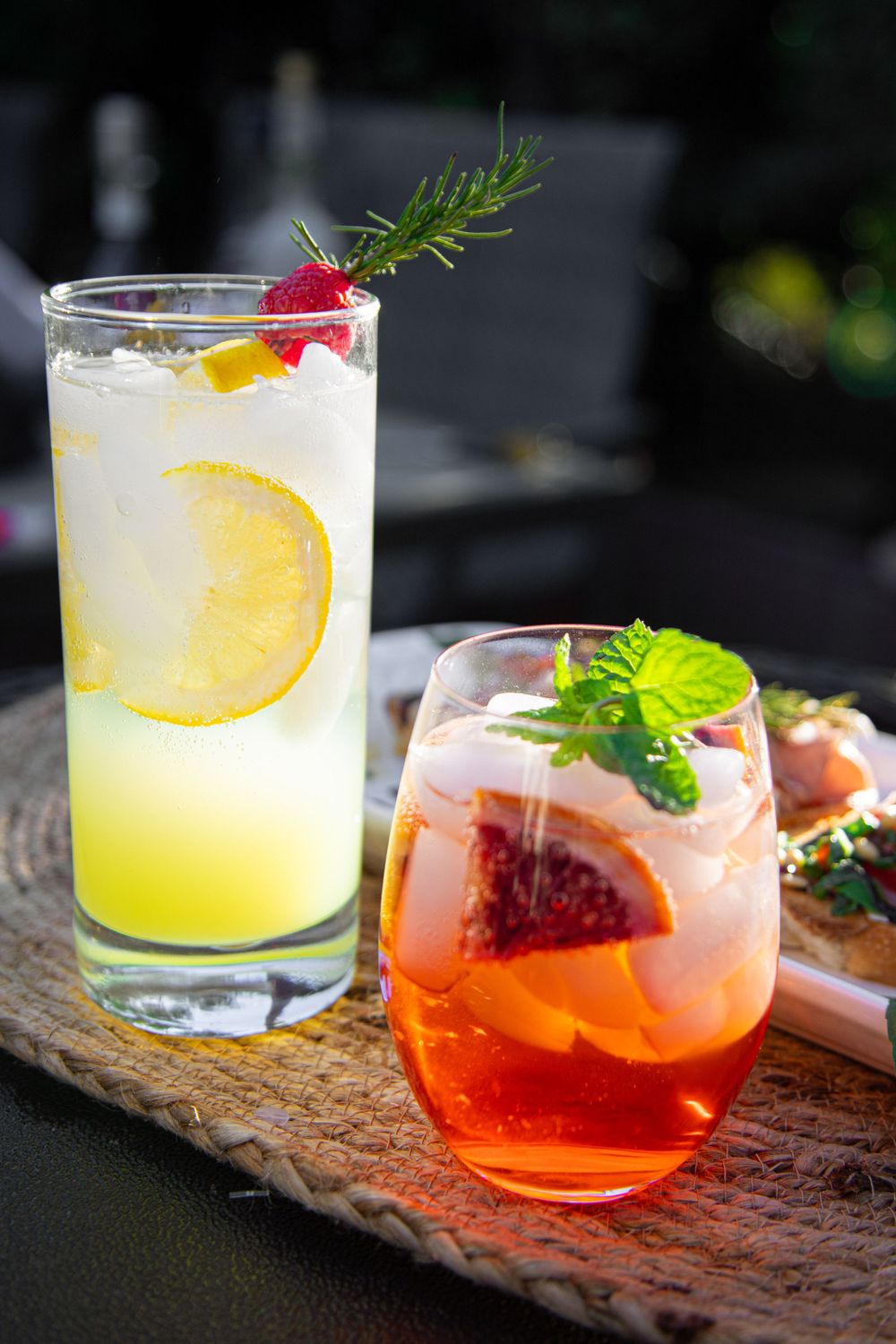 Two refreshing cocktails garnished with citrus, berries and herbs on a wooden board.