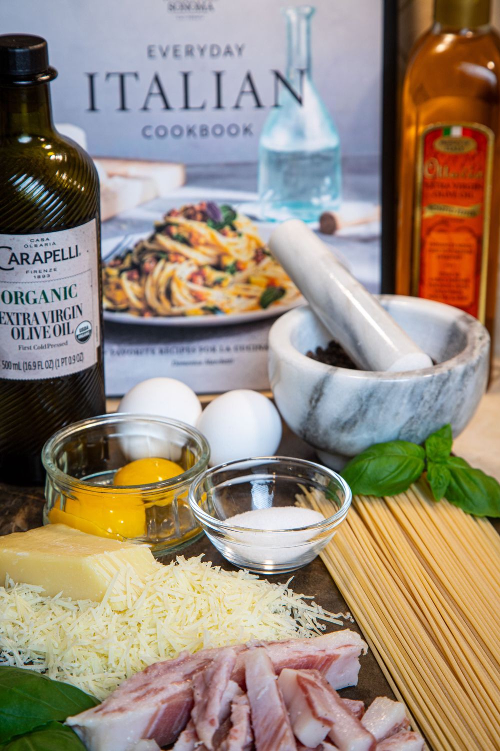 Italian cooking ingredients for carbonara with pasta, guanciale, olive oil, cheese and cookbook on marble counter.