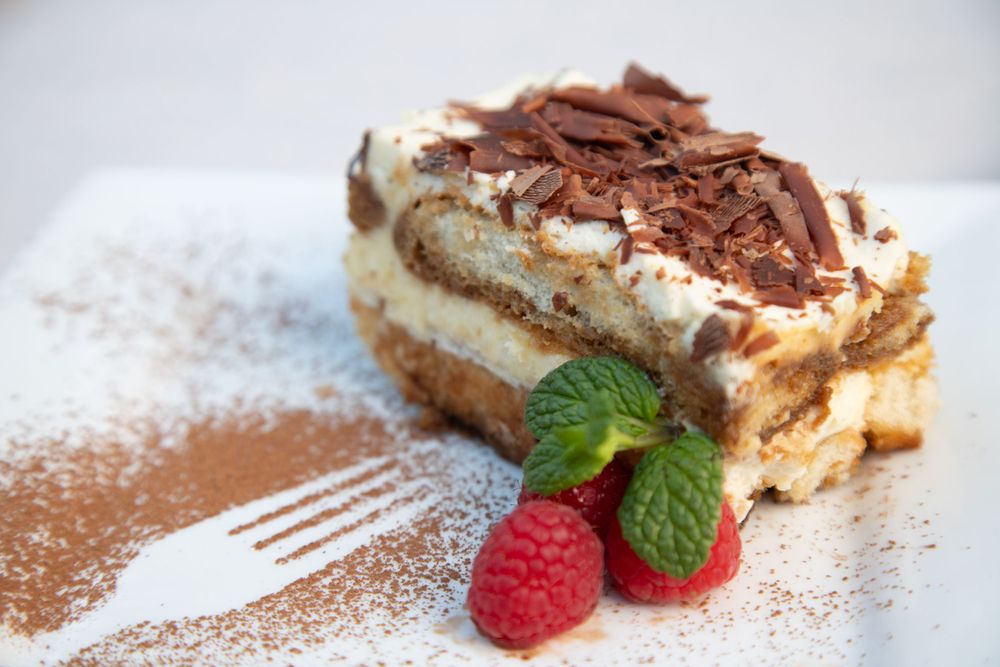 Layered tiramisu dessert topped with chocolate shavings, powdered cocoa, and fresh raspberries.