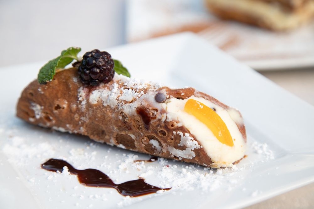 Chocolate cannoli filled with cream and topped with berries and mint on a white plate.