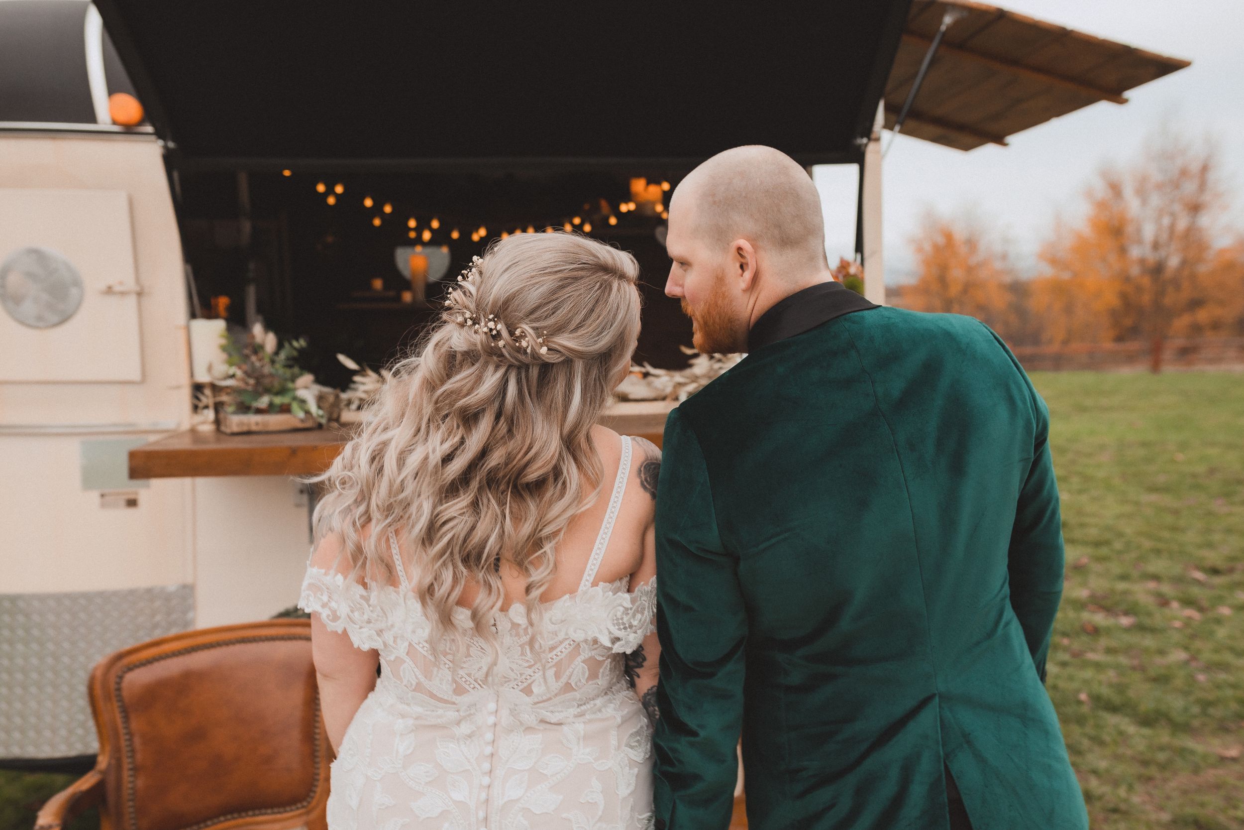 A couple in wedding attire standing at a food truck counter with fall foliage and string lights in the background.