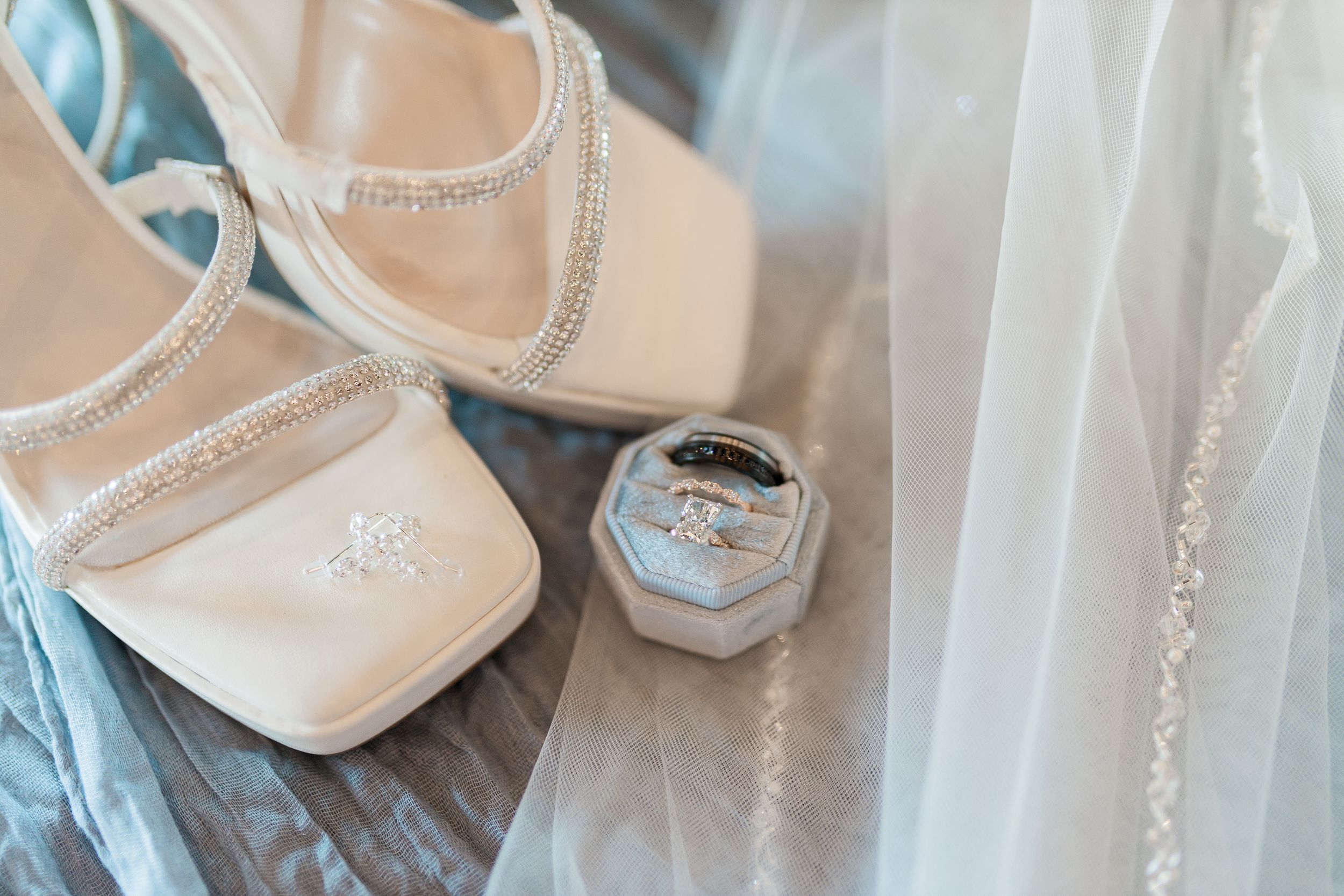 Elegant bridal shoes and platinum wedding rings displayed on delicate lace veil in soft lighting by Michigan wedding photography team.