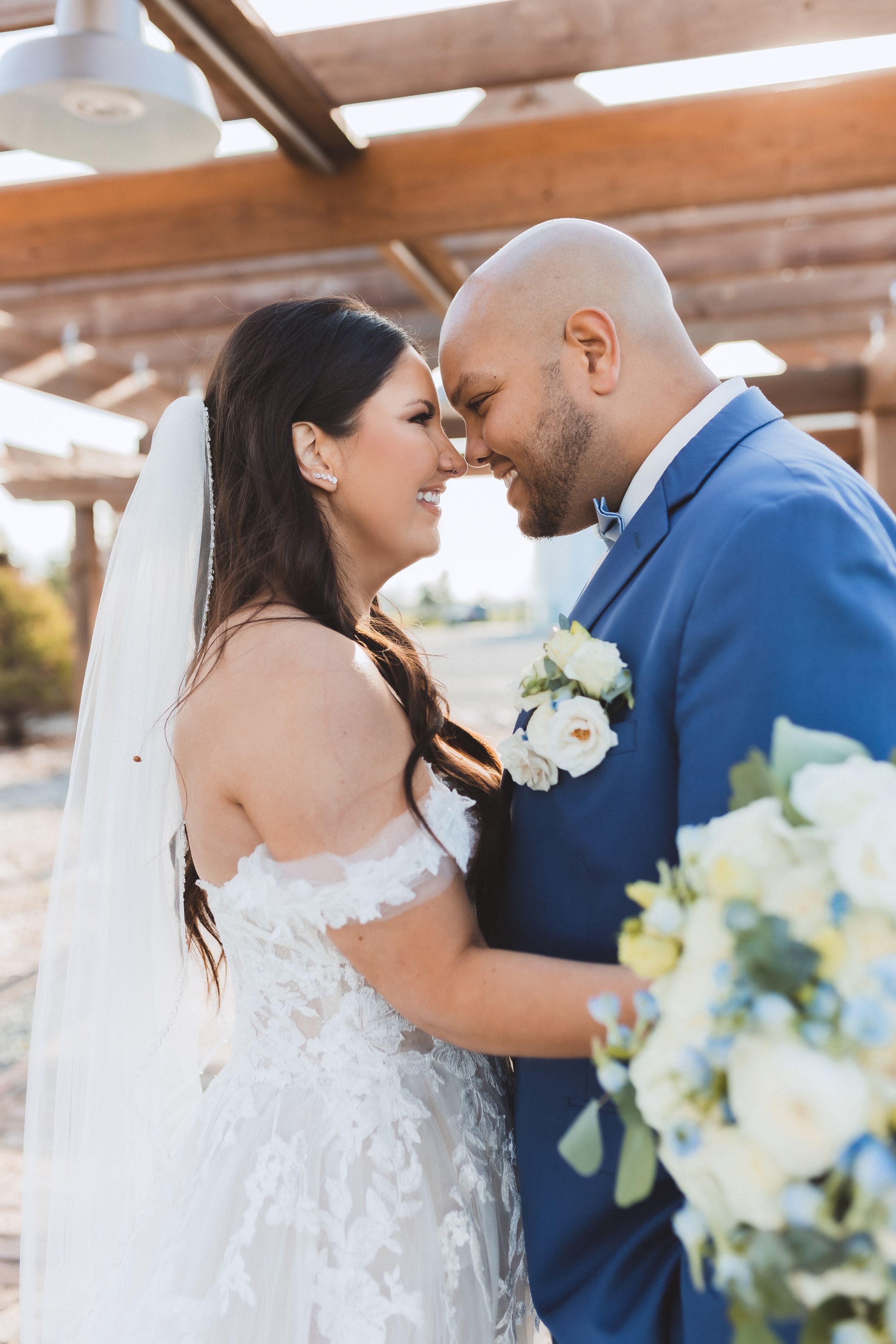 A happy couple shares an intimate moment on their wedding day under wooden beams with white flowers nearby.