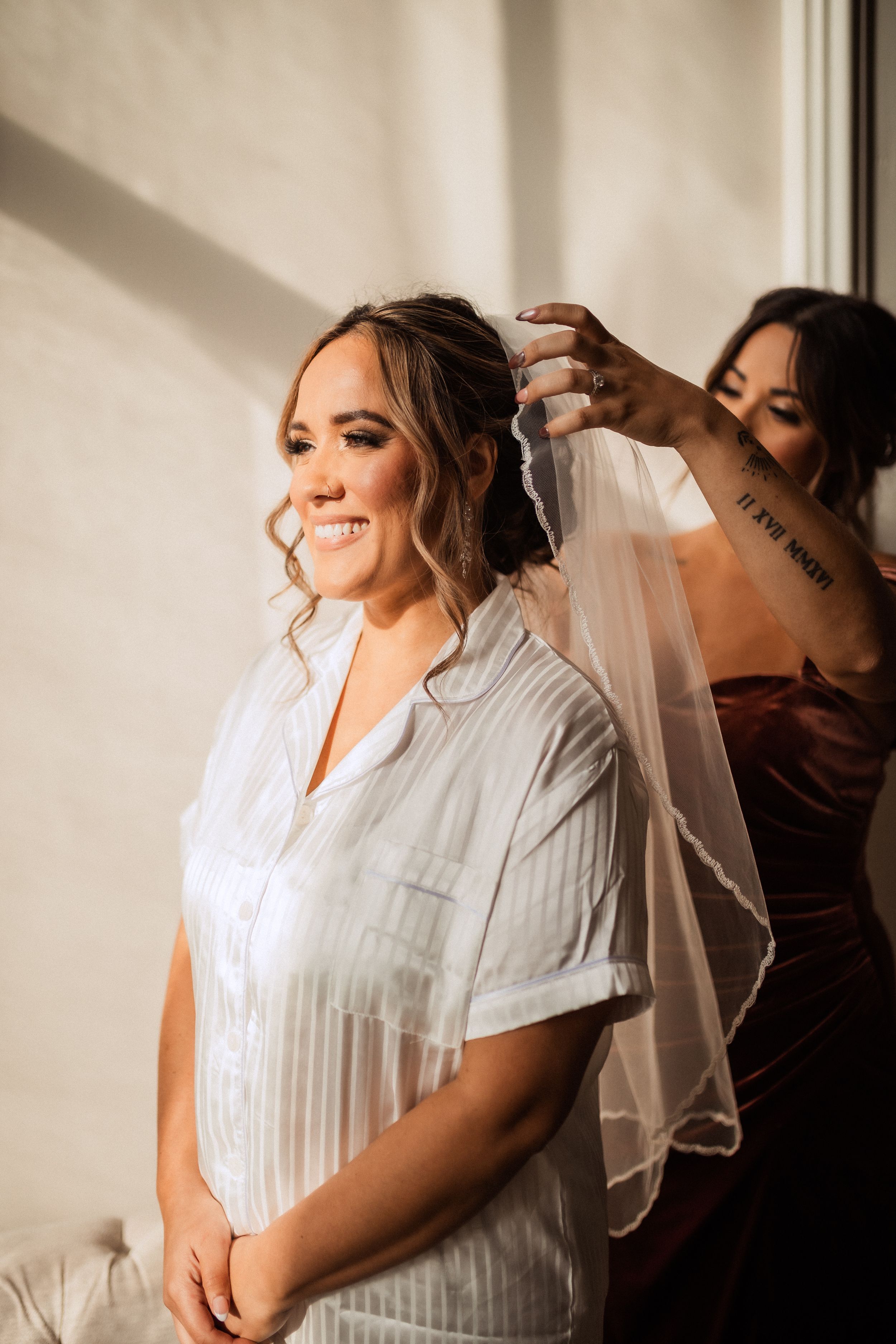 A person having their bridal veil placed while wearing a striped button-up shirt and smiling during wedding preparations.
