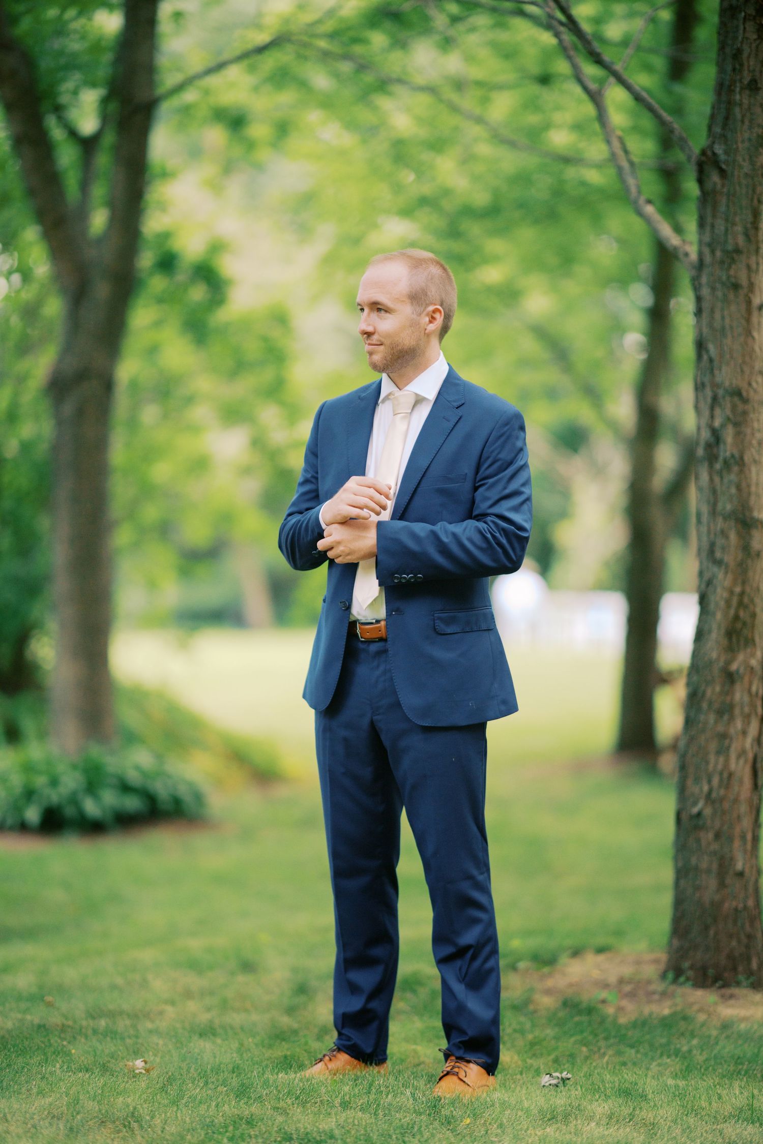 Person in a navy blue suit and light pink shirt stands thoughtfully in a park setting with green trees in the background.