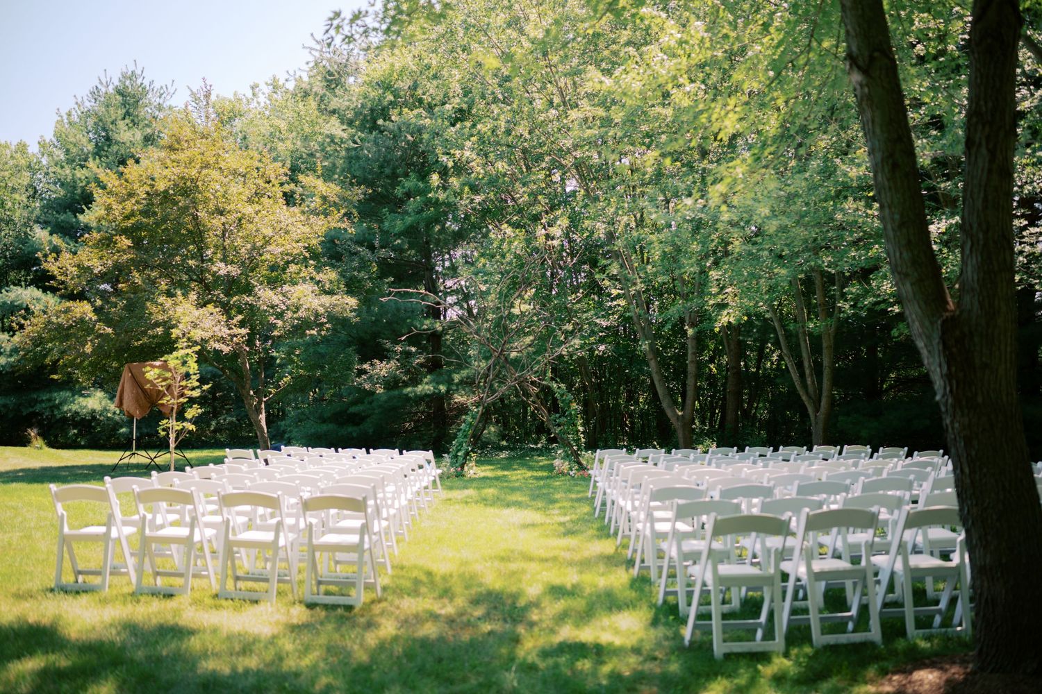 White folding chairs arranged in rows on a sunlit lawn surrounded by lush green trees for an outdoor wedding ceremony.