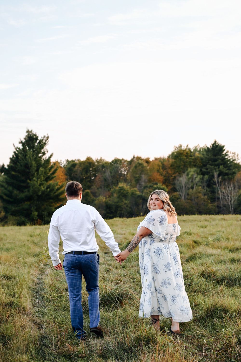 A couple walks hand-in-hand through a grassy meadow on an autumn day, the woman wearing a flowy floral dress.