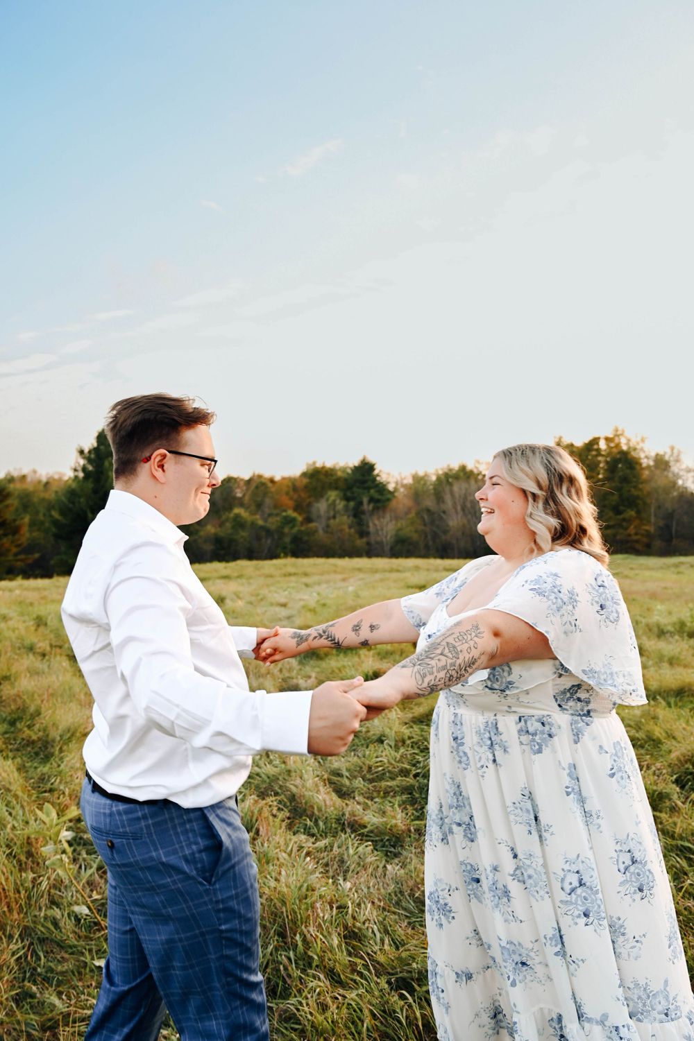 A romantic couple in a field share a joyful moment, with her wearing a floral dress and him in a white shirt.