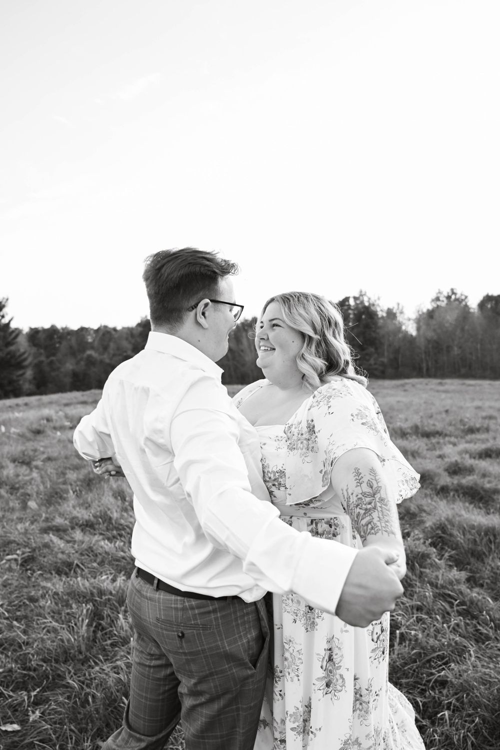 A black and white photo captures a couple dancing and laughing together in a meadow at sunset.