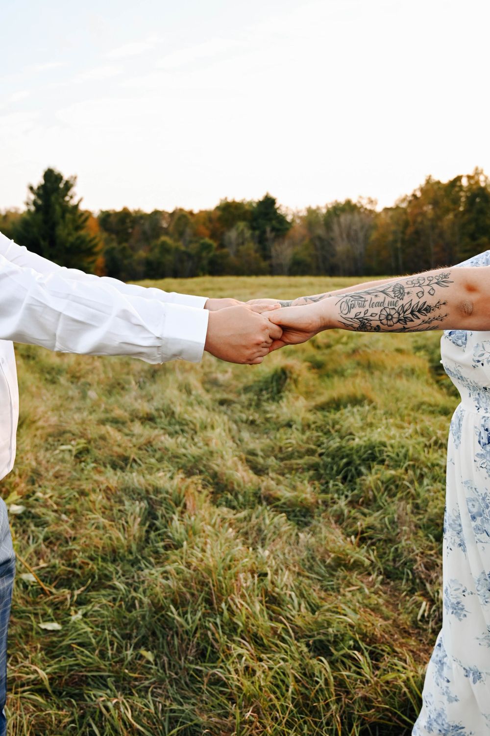 Two people holding hands in a grassy field with tattoo on one arm and long grasses at sunset.