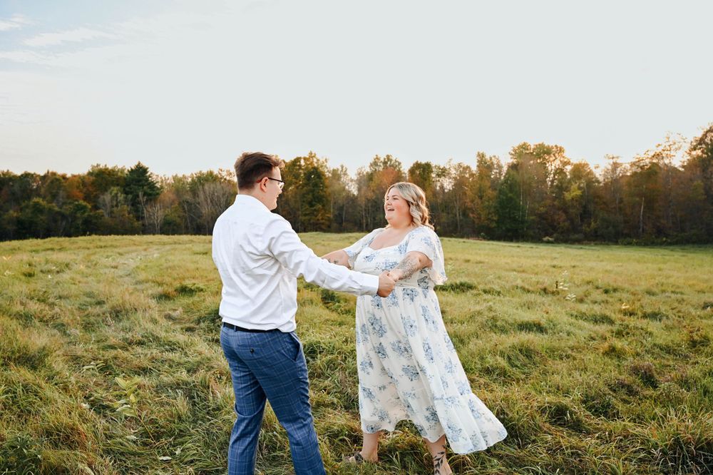 A couple dances together in a sunlit meadow during sunset with autumn trees in the background.