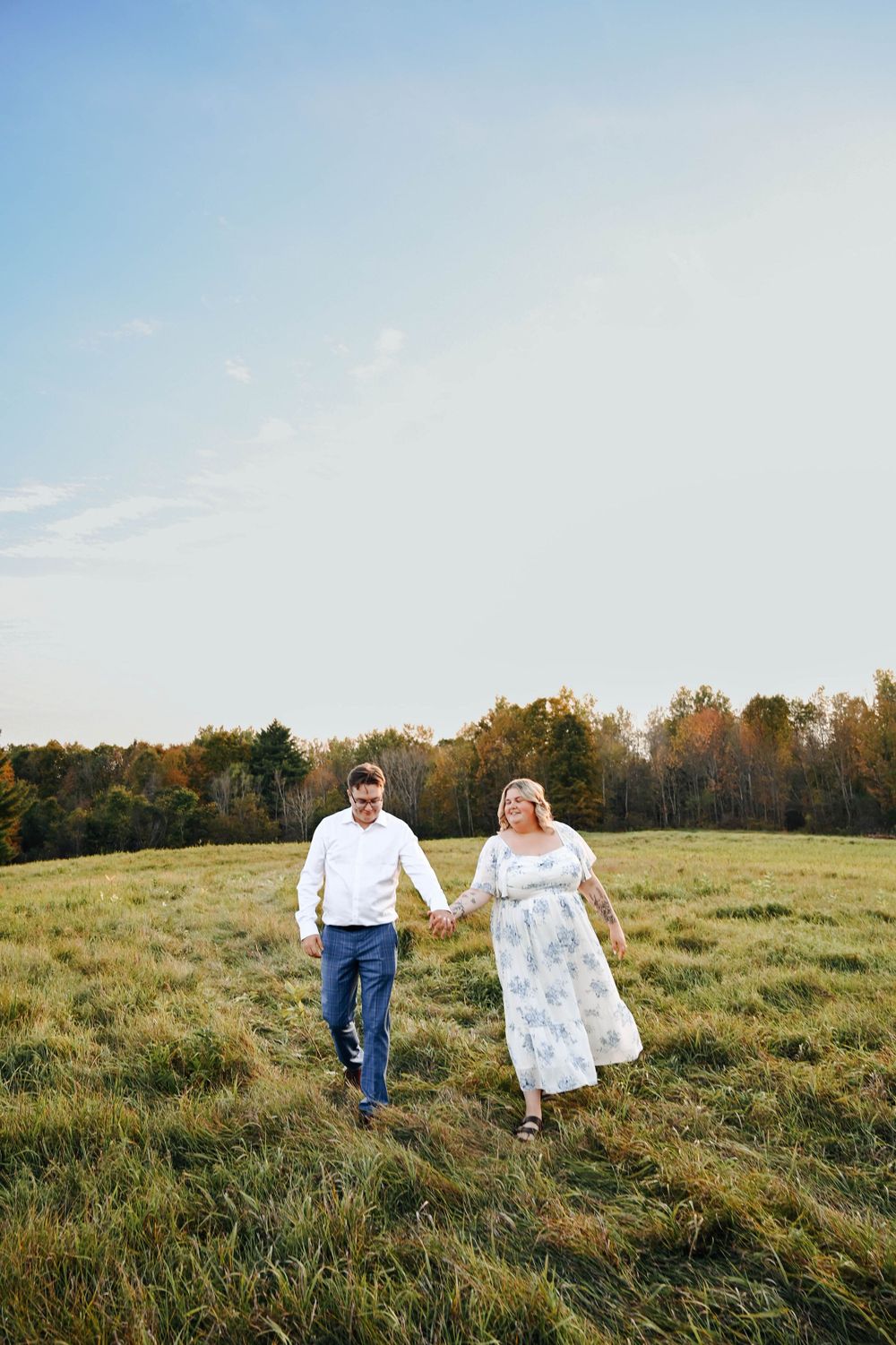A couple joyfully runs through a grassy meadow at golden hour with autumn trees in the background.