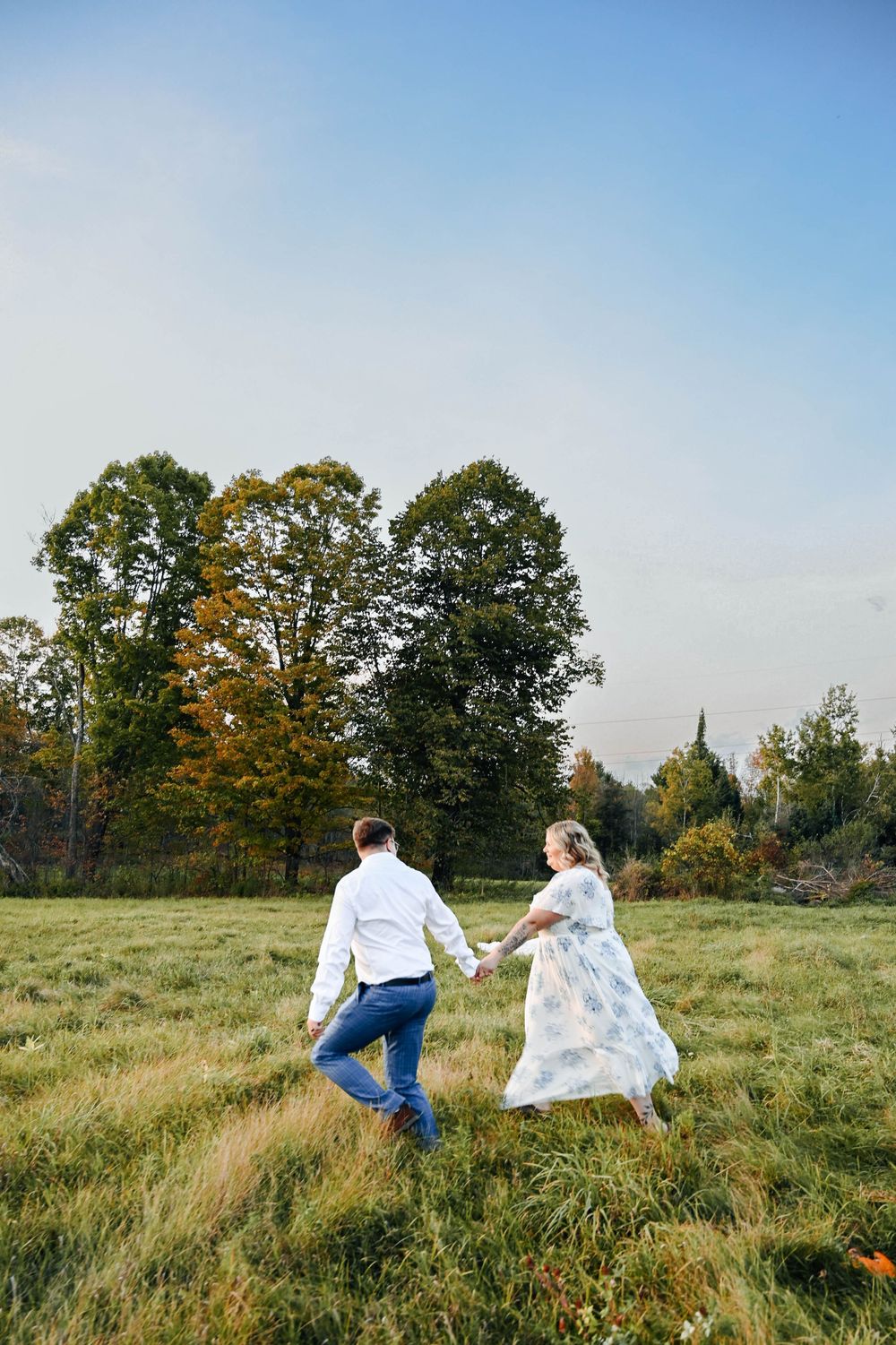 Two people in white outfits hold hands while running through a grassy field surrounded by autumn trees.