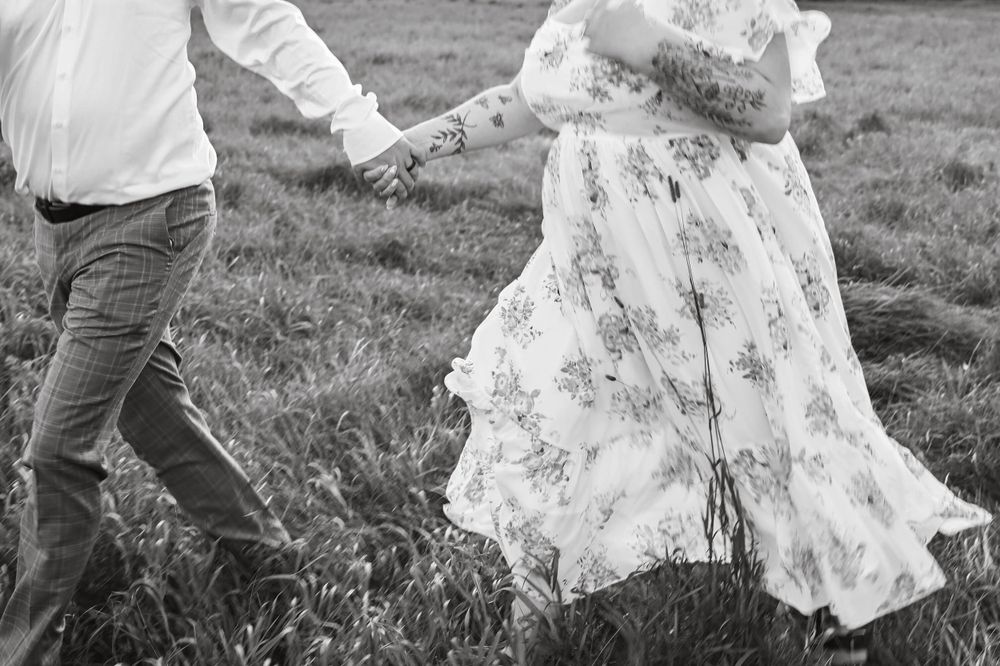Black and white photo of hands clasped together while walking through a field, with flowing floral dress details.