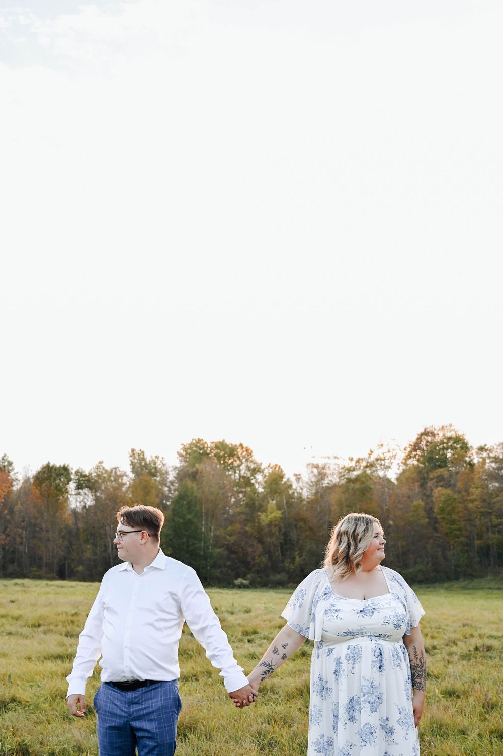 Couple holding hands while walking through a scenic autumn field with trees in the background on a sunny day.