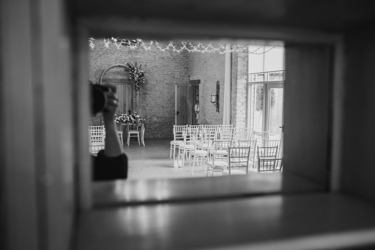 Black and white view through a window showing an elegantly decorated wedding ceremony space with white chairs.