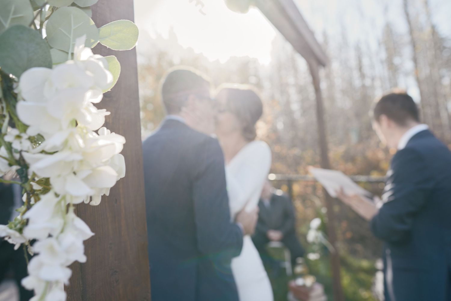 Bride & groom share a romantic kiss under an arch in golden autumn light, framed by white florals and a heartfelt backyard wedding ceremony.