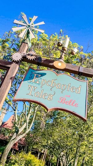 A wooden sign for 'Enchanted Tales Belle' with decorative windmill and gears against a bright blue sky in a theme park setting.
