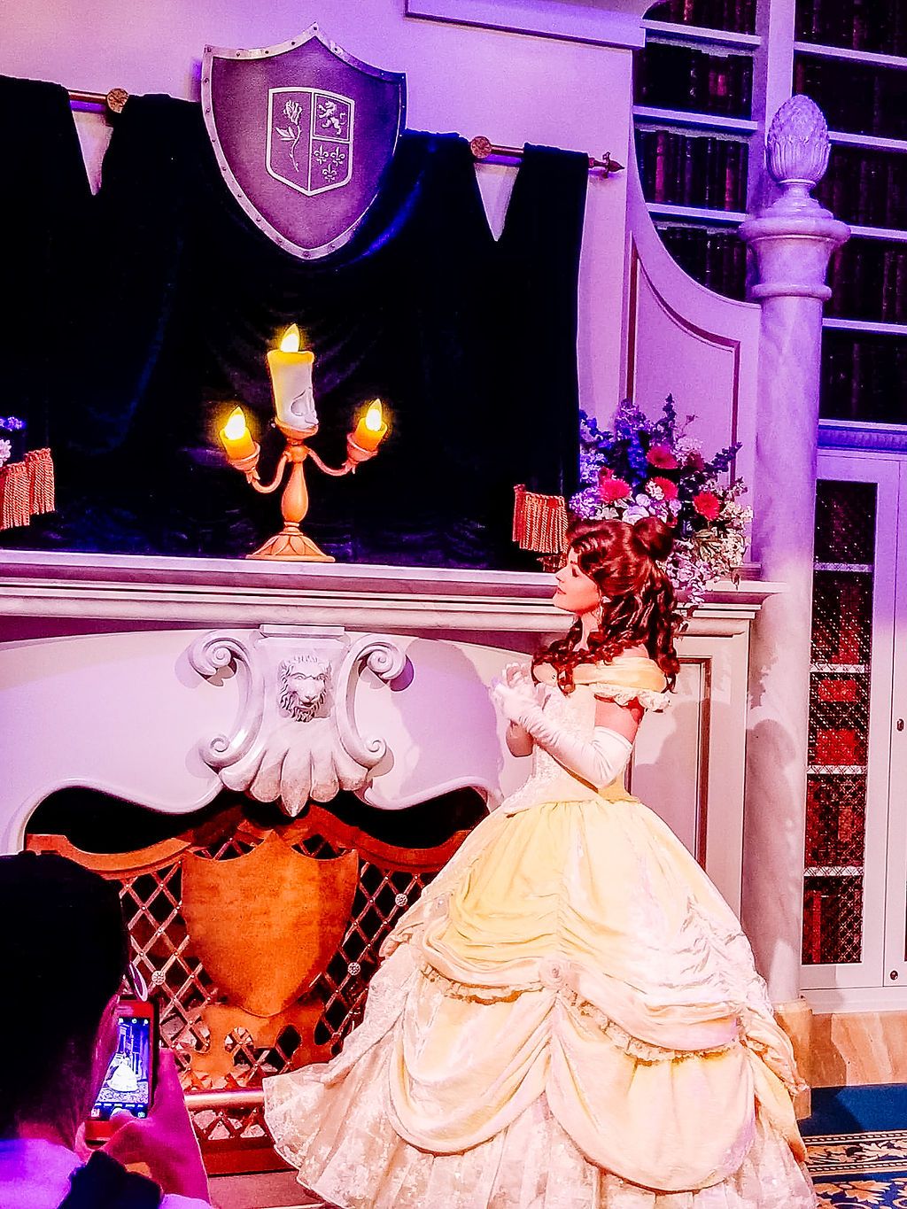 An ornate ballroom scene with a yellow gown against purple lighting, featuring a shield crest and candlestick decor.