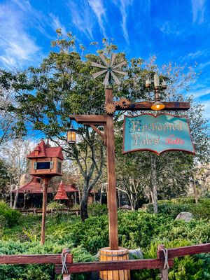 A wooden sign for Enchanted Tales with a colorful birdhouse and lush trees under vibrant blue skies at a theme park.