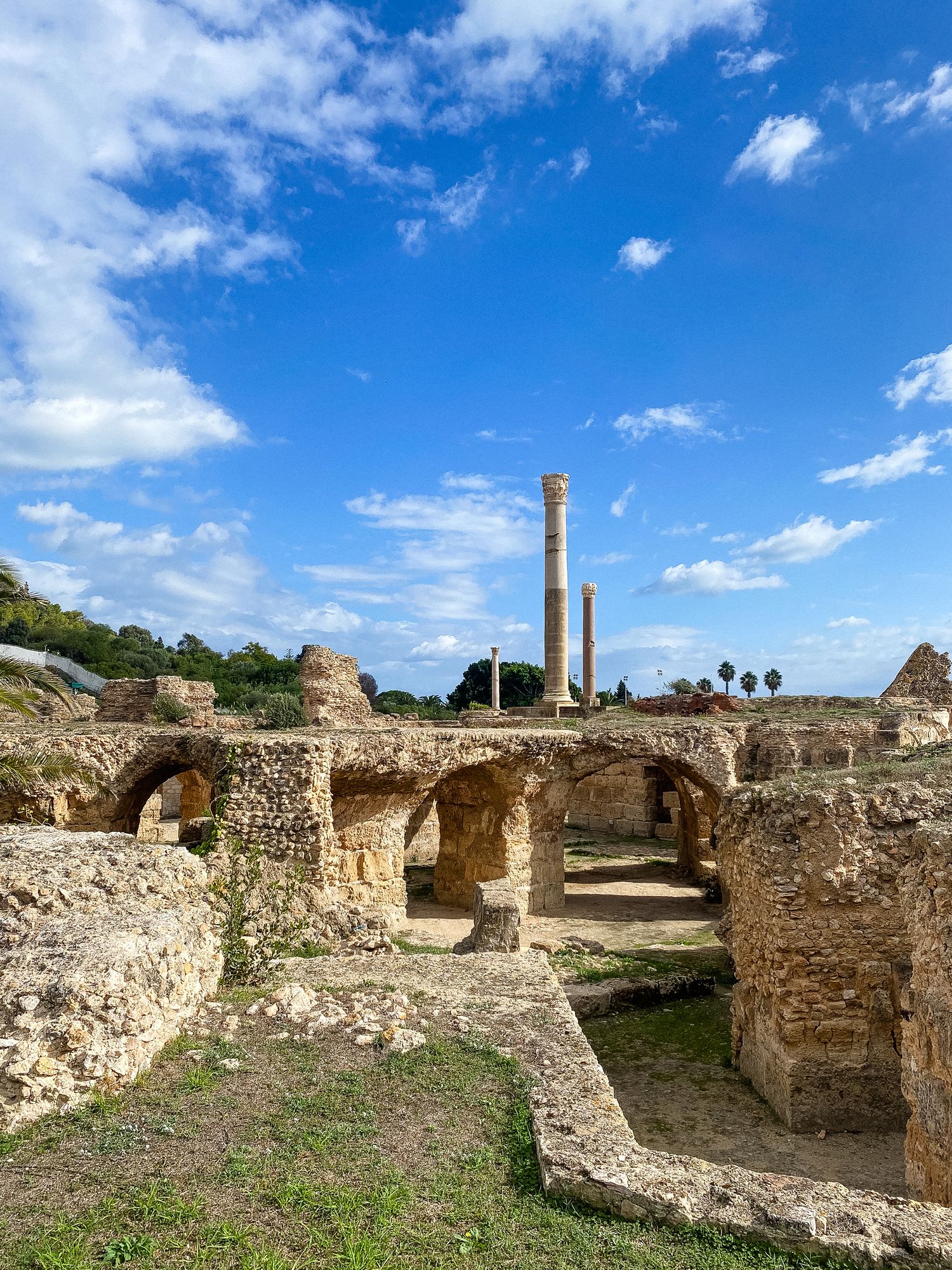 Ancient Roman ruins with stone arches and columns at Carthage Antonine Baths against bright blue sky with fluffy clouds.