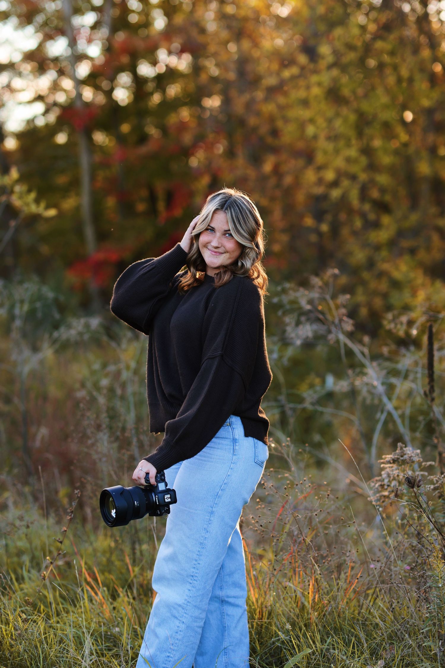 Relaxed portrait against white backdrop features denim overalls paired with cream sweater while holding camera.