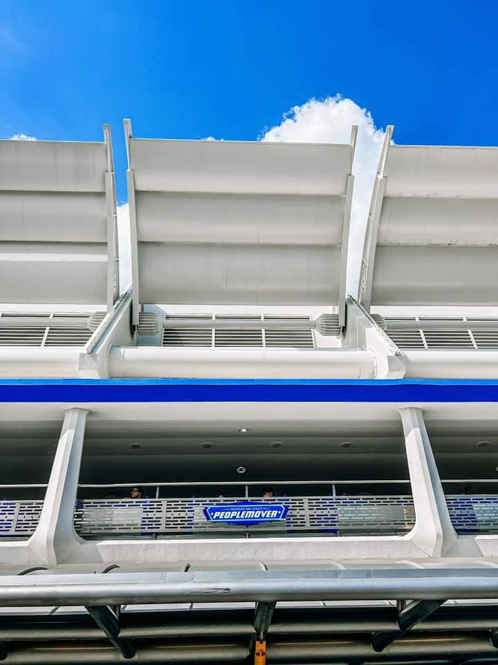 Modern train station platform with white angled ceiling panels and blue sky visible through gaps on a sunny day.