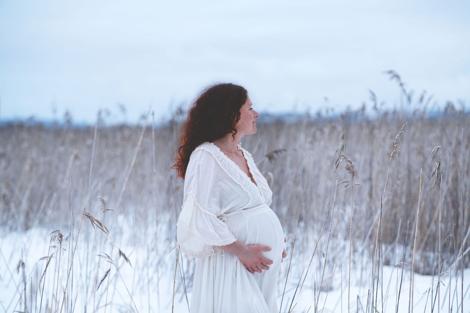 Gravid rödhårig kvinna i vit klänning som står i vassen vid en sjö. Det är en mulen vinterdag med snö.