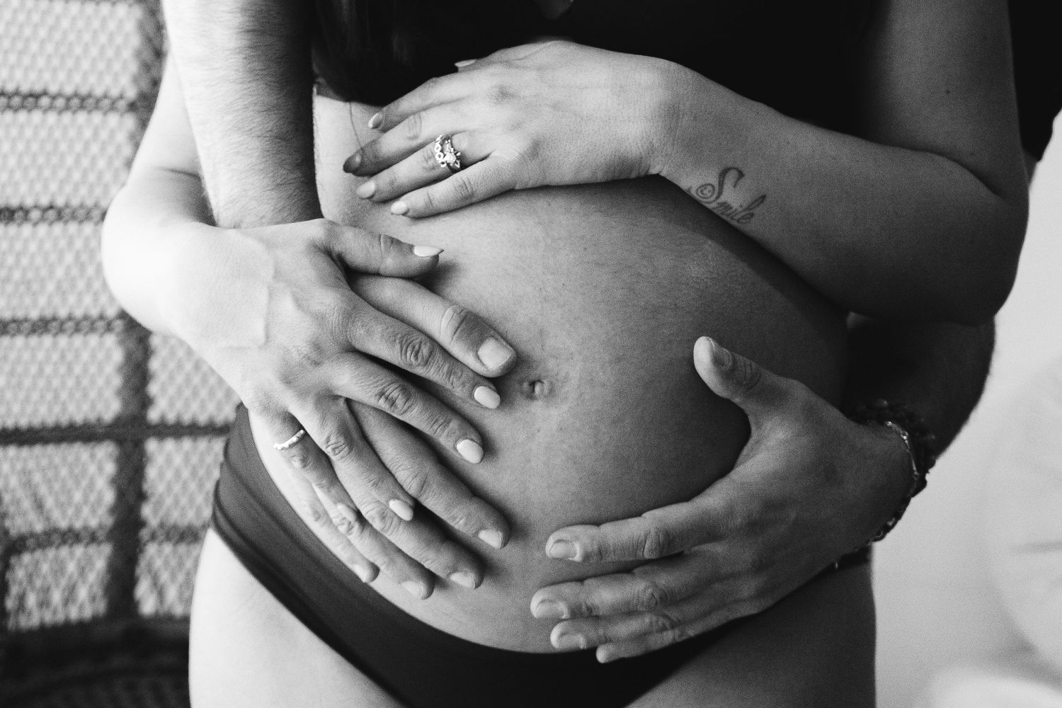 Foto in bianco e nero di mani che cullano la pancia incinta con tatuaggio visibile.