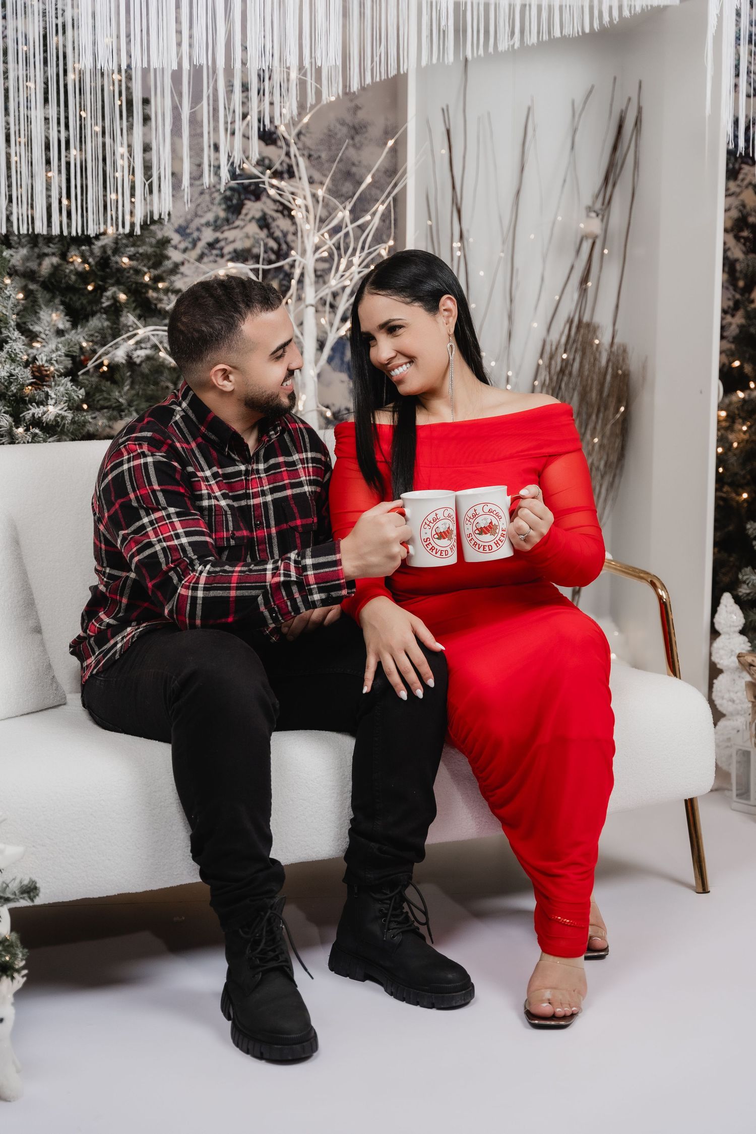 A couple sits on a white sofa during a holiday photoshoot, with the woman in red and man in plaid sharing a festive moment.