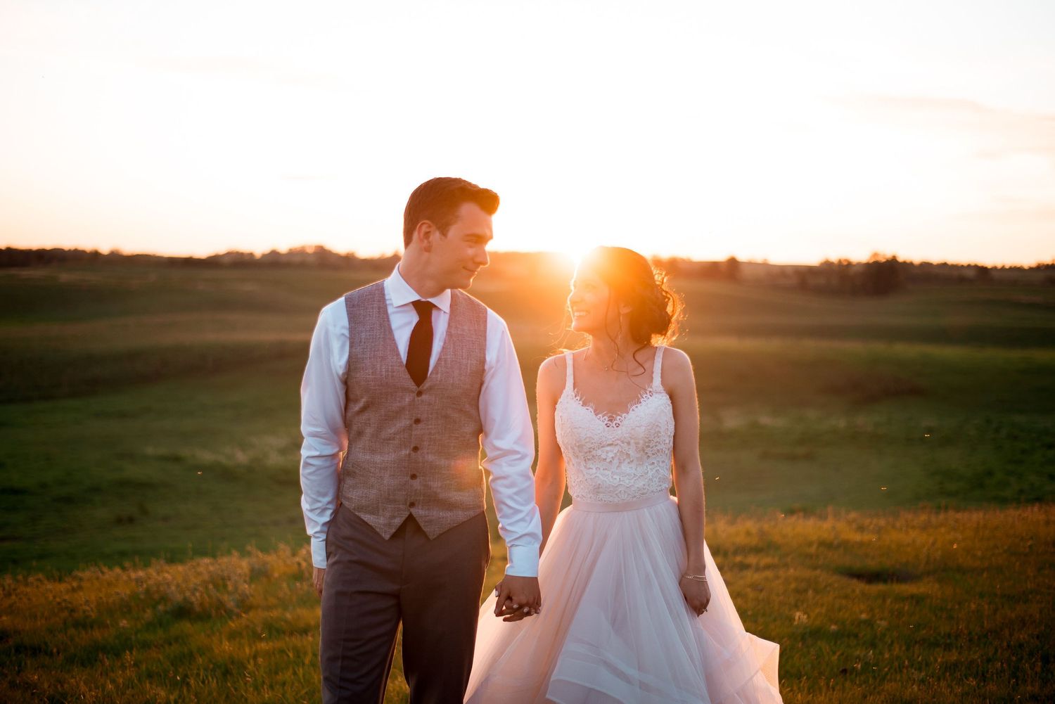 A couple in wedding attire stands together in a grassy field at sunset with golden sunlight behind them.