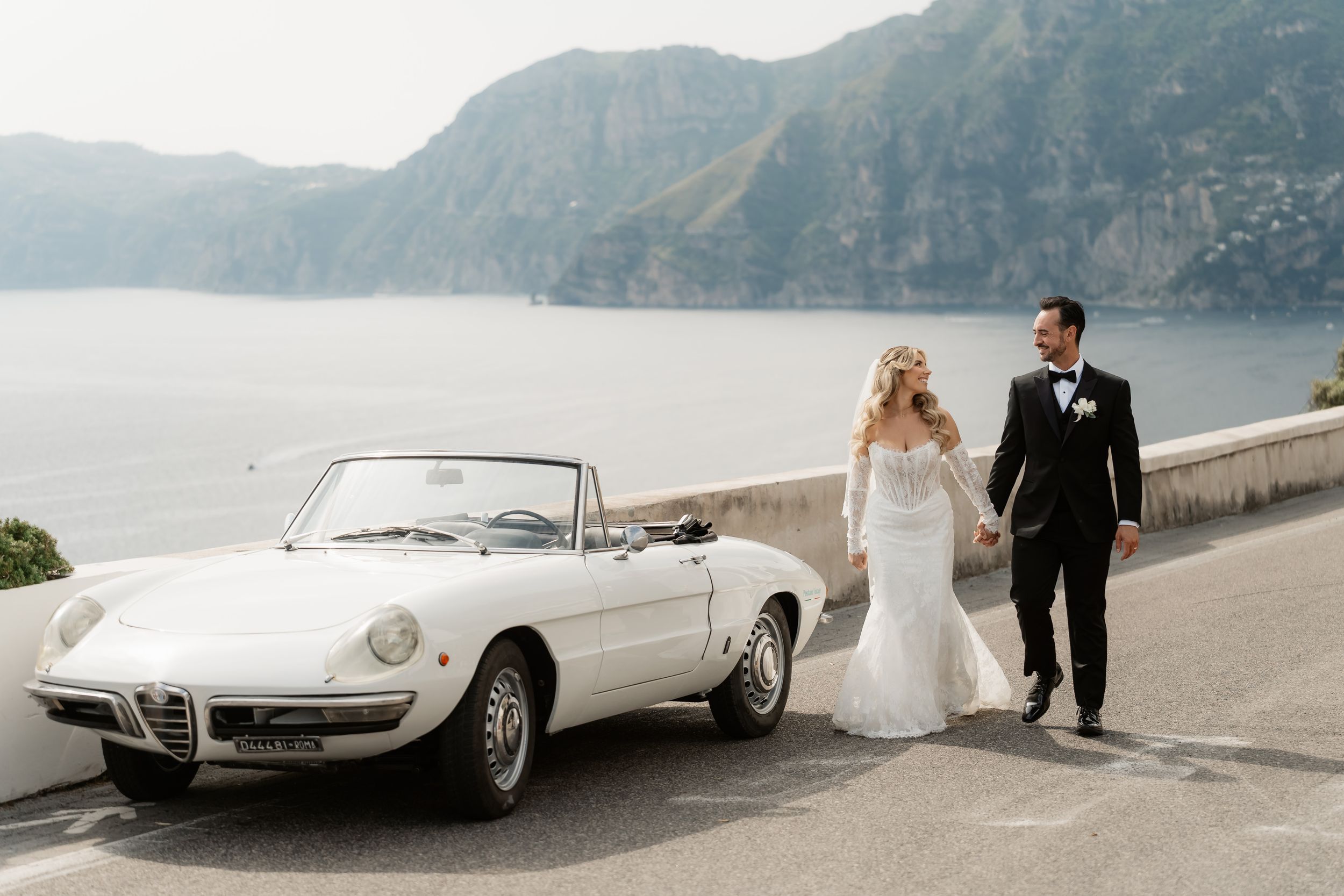 Bride and groom walk beside a vintage white Alfa Romeo Spider convertible with stunning Amalfi Coast cliffs and sea views.