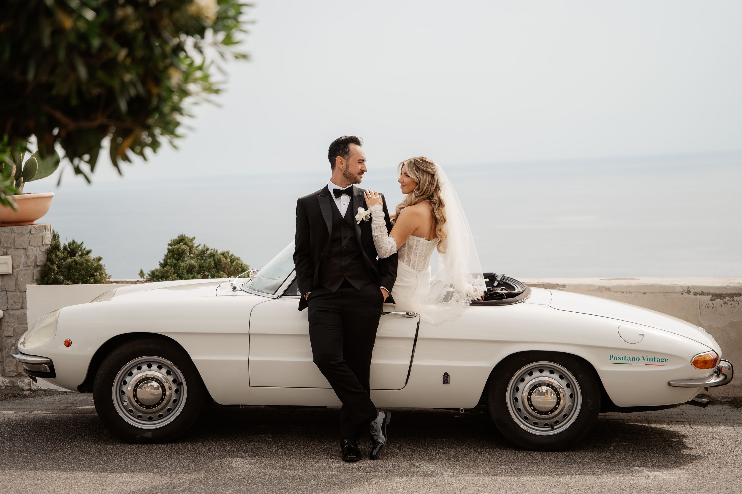Bride and groom pose beside a classic white convertible car with ocean views in the background on their wedding day.