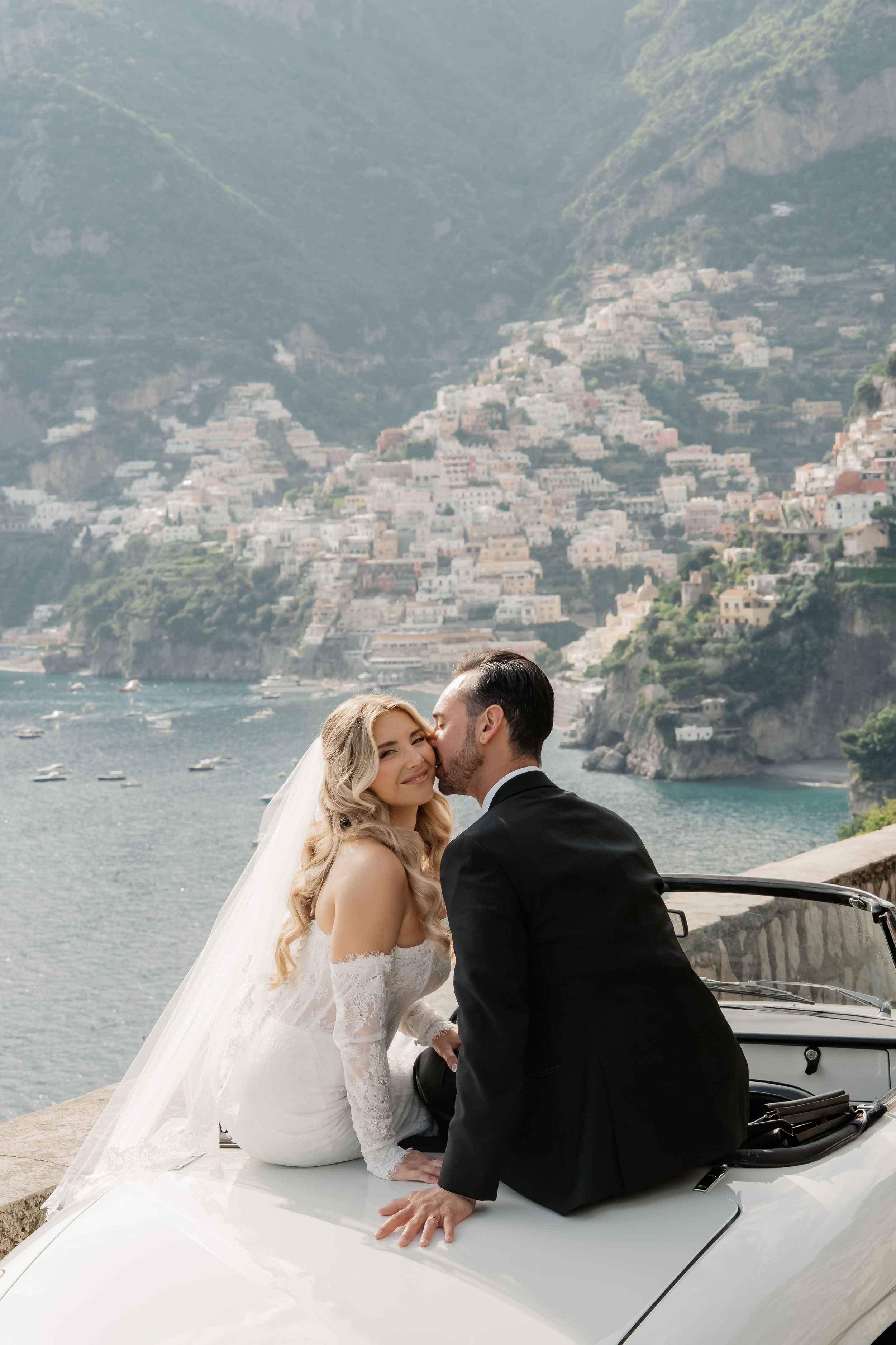Bride and groom sit on a white convertible car with Positano, Italy cliffside village and blue sea in the background.