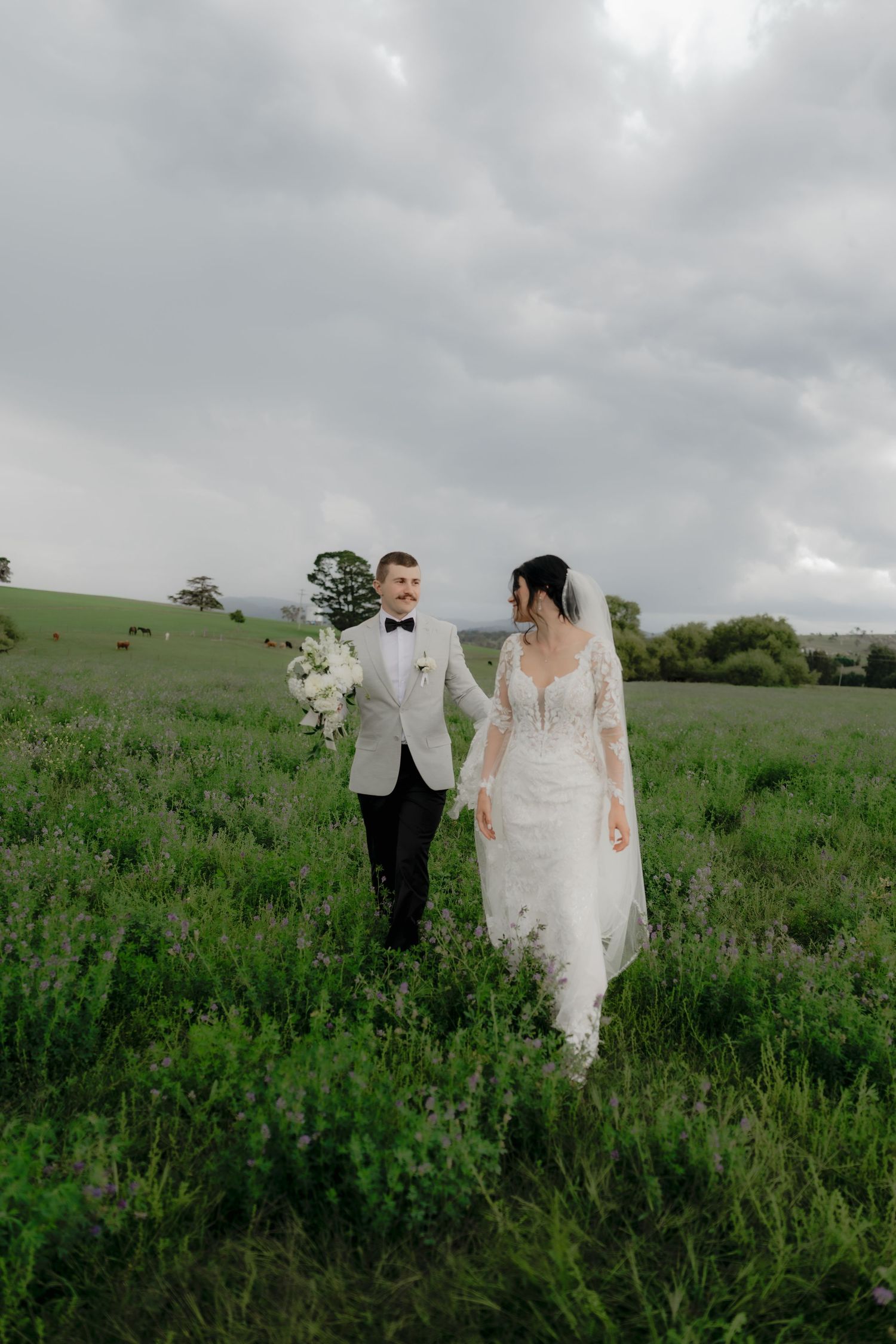 Bride and groom walk hand in hand through lush green lavender field under cloudy sky on their wedding day.