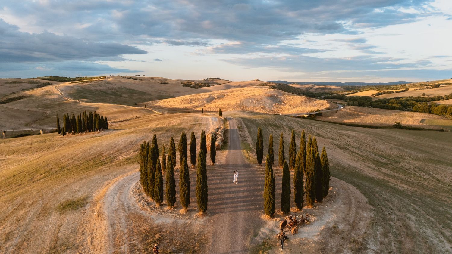 Aerial view of a Tuscan chapel surrounded by cypress trees on a winding dirt road amid golden rolling hills.
