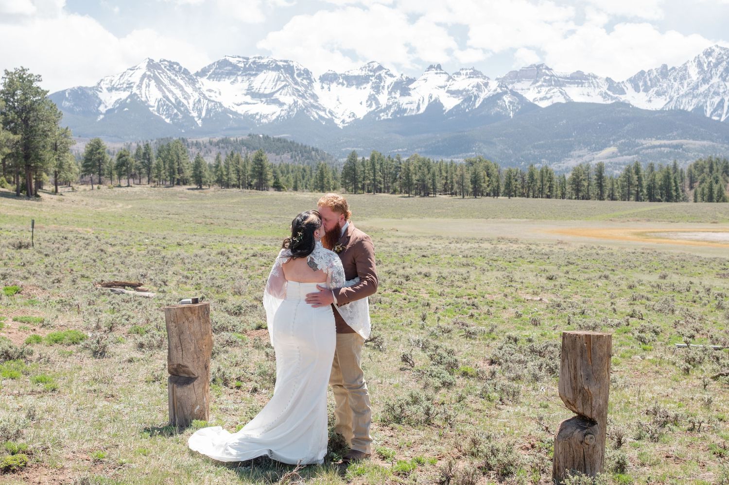 Bride and groom share a romantic moment in a mountain meadow with snow-capped peaks and pine trees in the background.