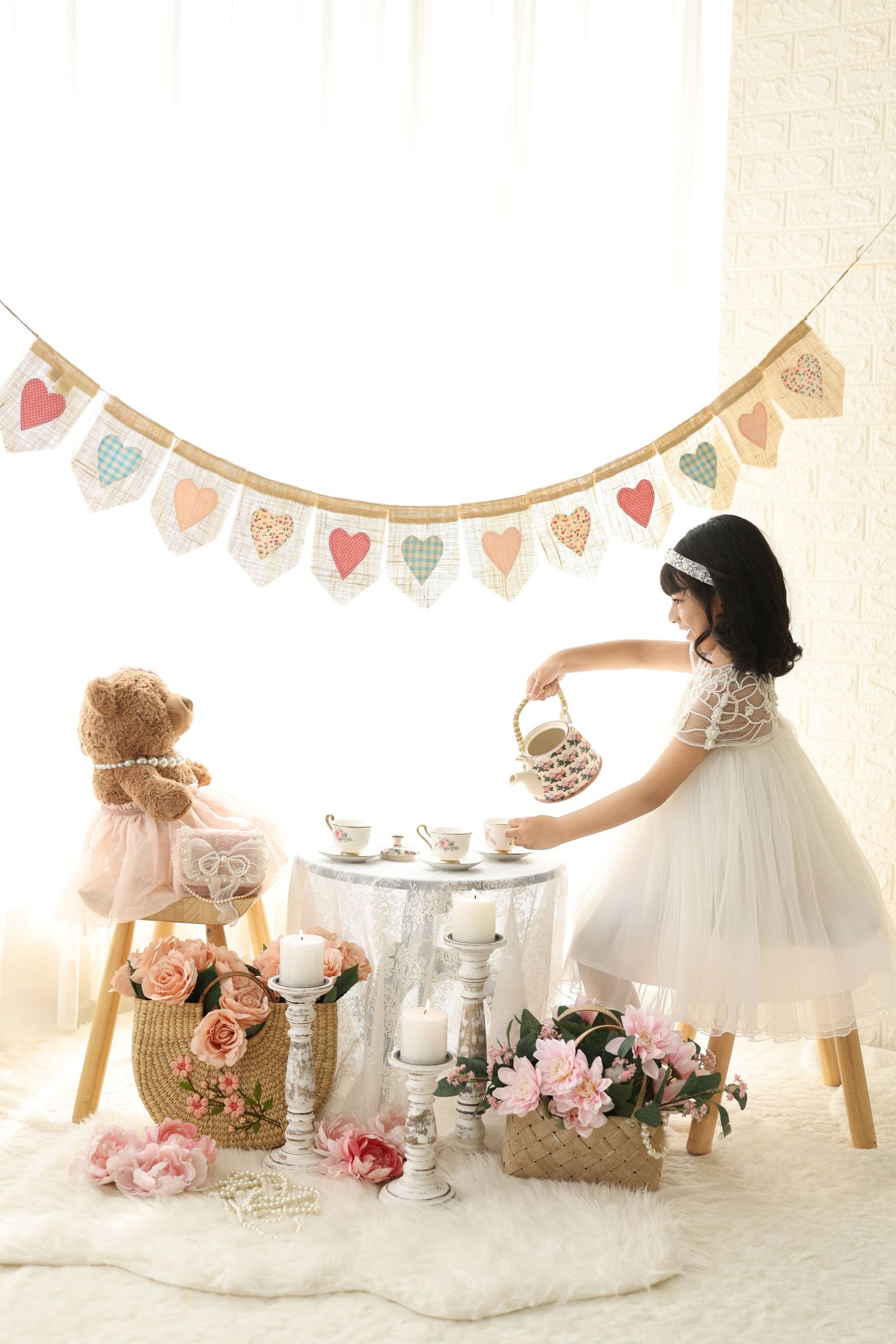 Young girl in white dress hosting a teddy bear tea party with flowers, candles, and heart bunting decorations.