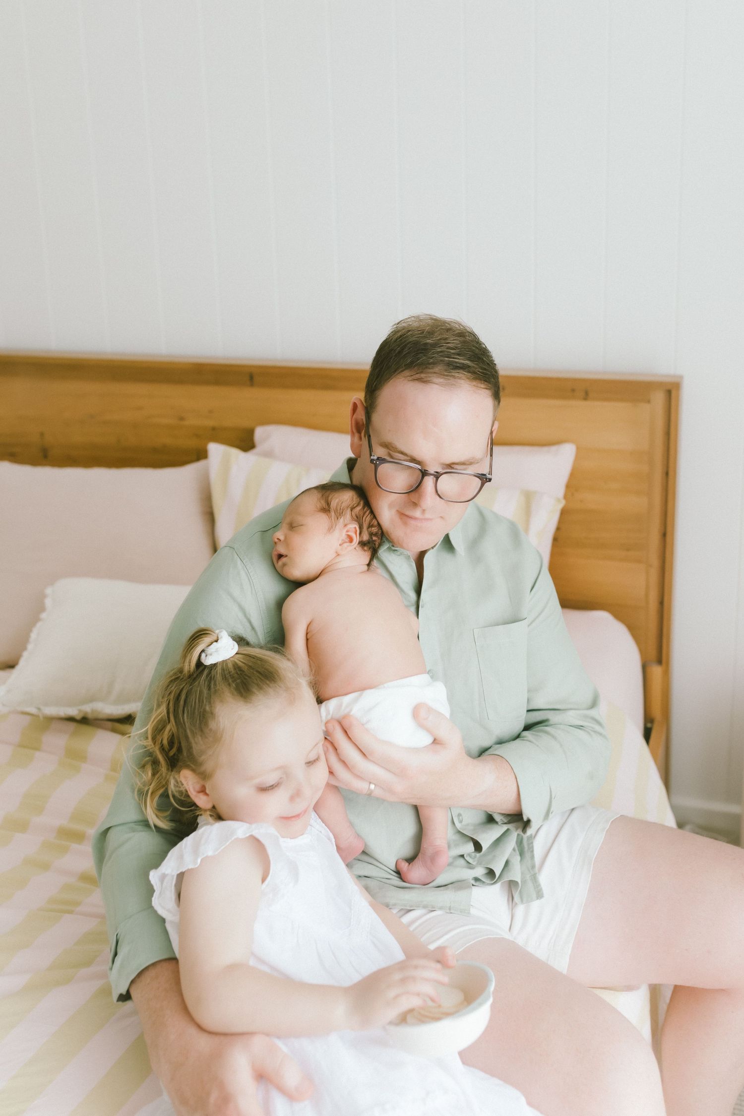 Parent in green shirt holds newborn baby while young toddler girl in white dress looks on, sitting on bed.