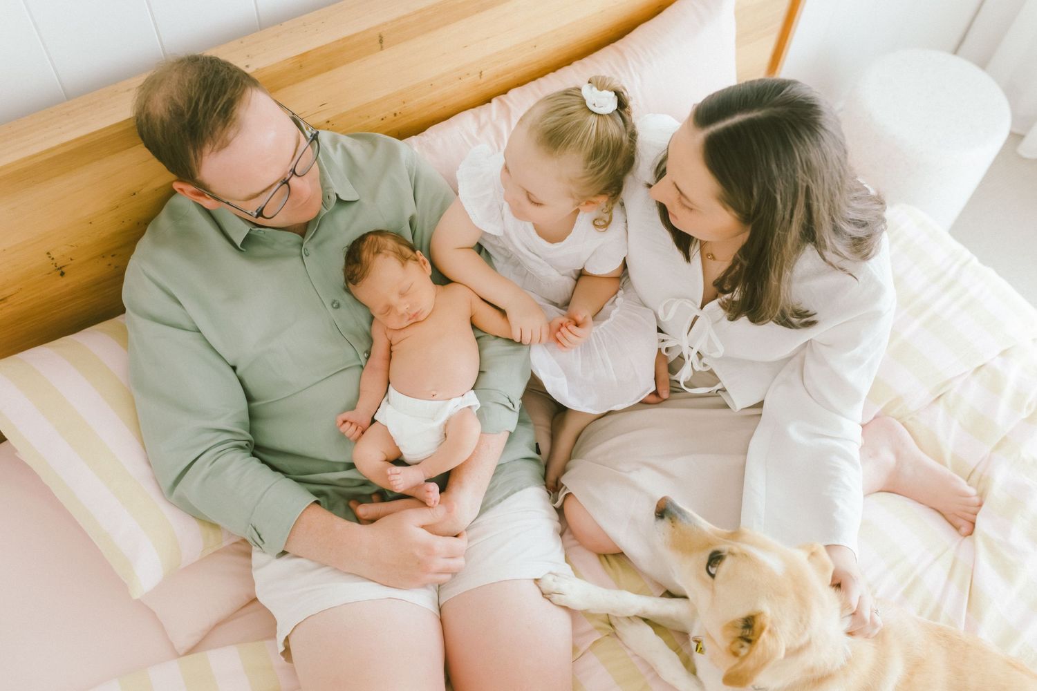 Family with newborn baby, toddler girl, and golden retriever dog sitting together on a bed in a bright room.